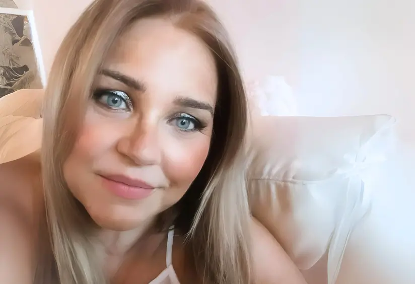 Microblading Ponferrada Una mujer de larga melena rubia y ojos azules sonríe a la cámara. Está en un interior, con una almohada blanca y una pared detrás. Sus cejas impecables, perfeccionadas con microblading en Ponferrada, complementan su camiseta blanca sin mangas. La imagen tiene un tono suave y cálido. La Orquídea Ponferrada